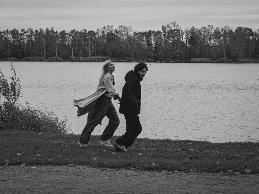 Black and white capture of a couple joyfully running by a lakeside in fall.