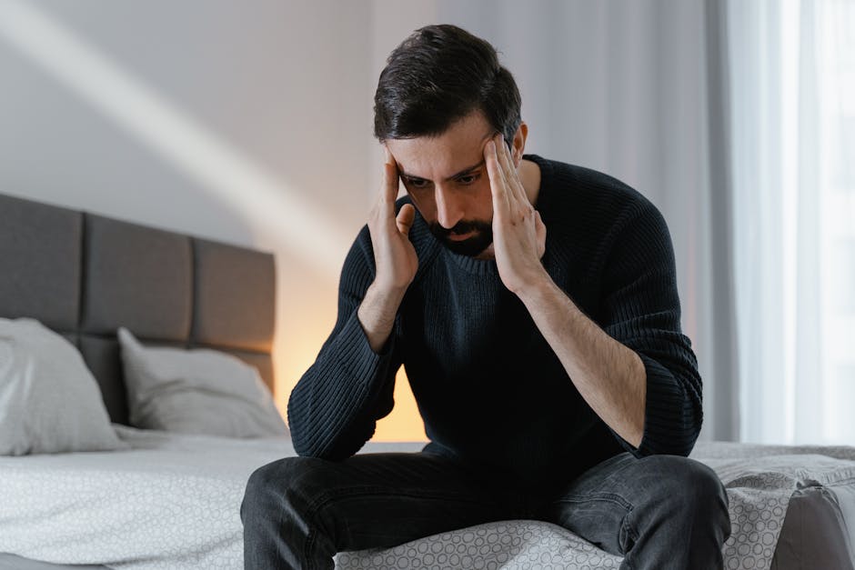 A man in a black sweater sits on a bed, expressing frustration in a bedroom.