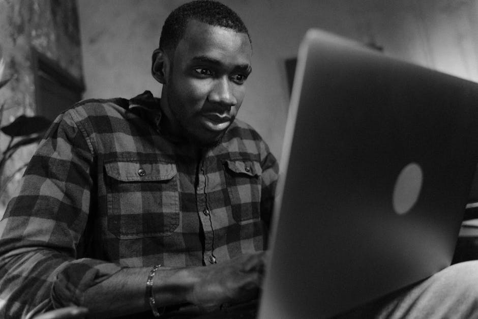 Black and white portrait of a man intently working on a laptop in a cozy indoor setting.