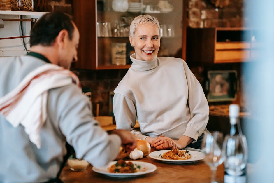 Smiling adult couple in casual clothes having dinner while male cooking pasta on plates at counter near glasses and wine bottle near various products and ingredients in light kitchen