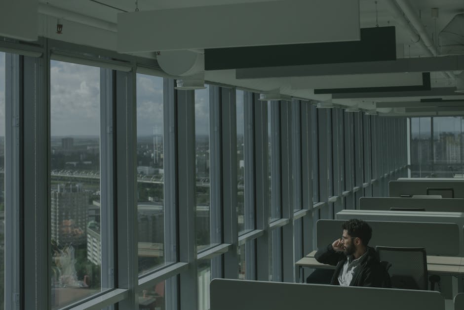 A man sits in a modern high-rise office, gazing thoughtfully through expansive windows at the city below.