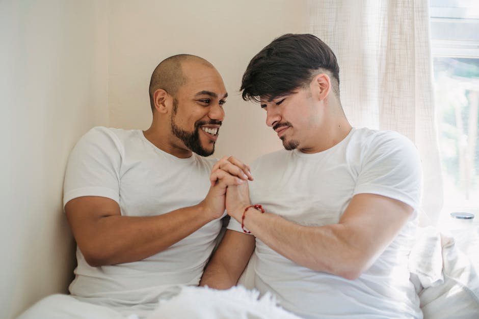 Two men sharing a moment of affection on a bed, holding hands and smiling naturally.