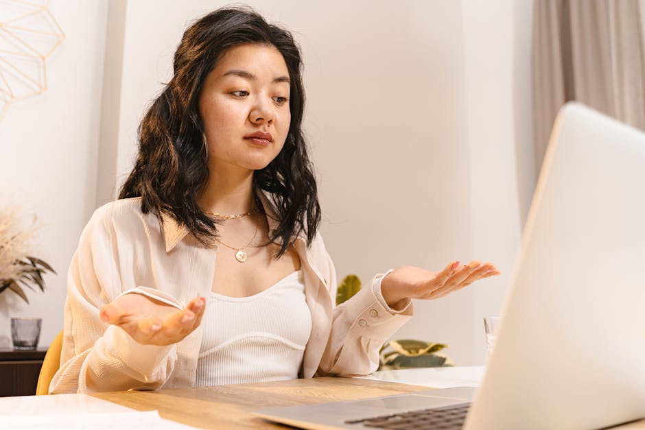 Asian woman in a long sleeve shirt, appearing puzzled while sitting indoors and looking at a laptop.