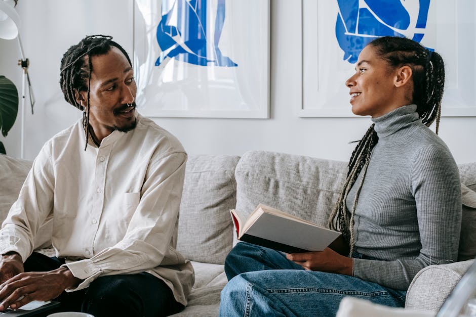 Positive young black lady with Afro braids sitting on sofa with interesting book in hands and speaking with boyfriend working remotely on laptop
