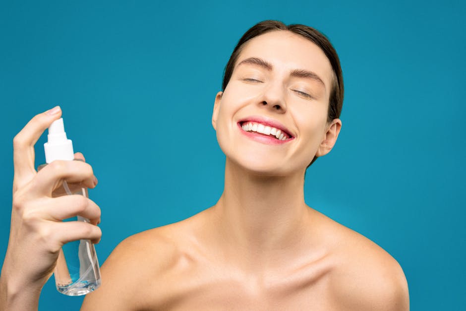 A joyful woman sprays facial mist with closed eyes against a blue background, showcasing skincare and happiness.