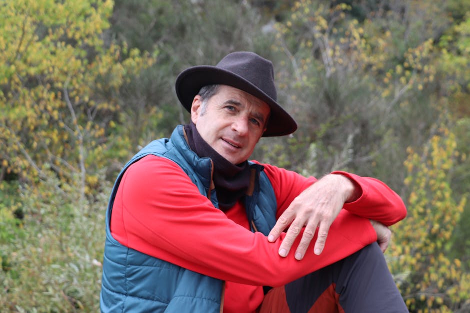 A middle-aged man wearing a fedora and orange jacket, posing outdoors in a natural setting.