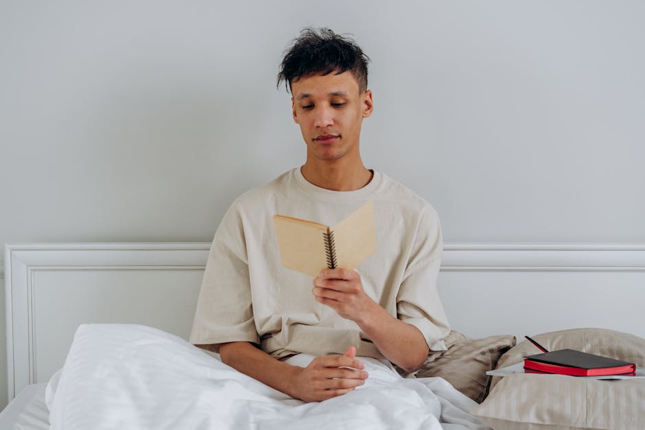 A young man sitting in bed reading with a notebook beside him, conveying relaxation and study.