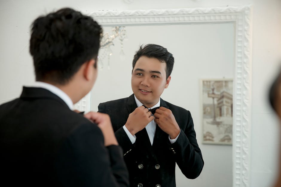 A man in a suit adjusts his bow tie while looking at his reflection in a mirror.