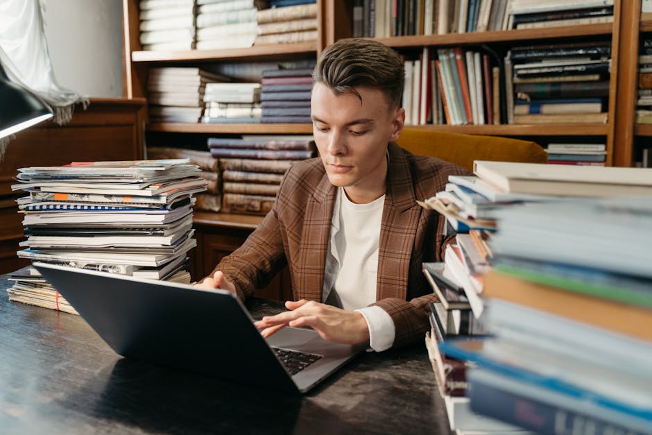 A focused young man studies on a laptop amidst stacks of books in a cozy library setting.